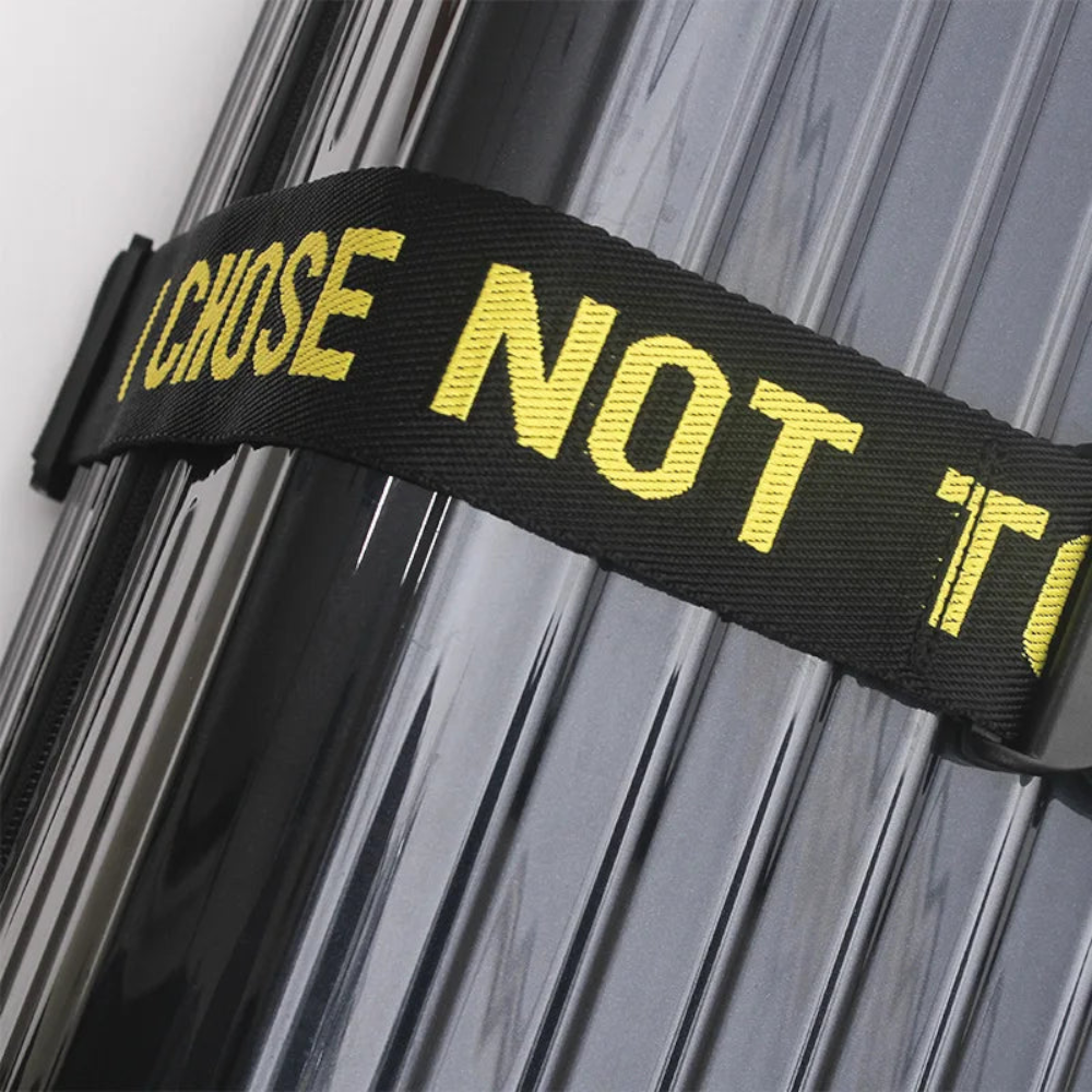 AïtaVia black luggage strap with yellow text "I CHOSE NOT TO" secures a grey ribbed suitcase.