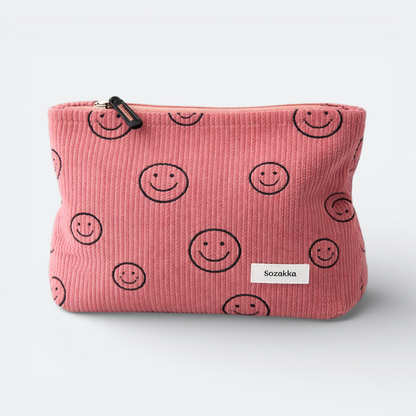 AïtaVia pink corduroy travel toiletry bag with a repeating smiley face pattern and a white label.